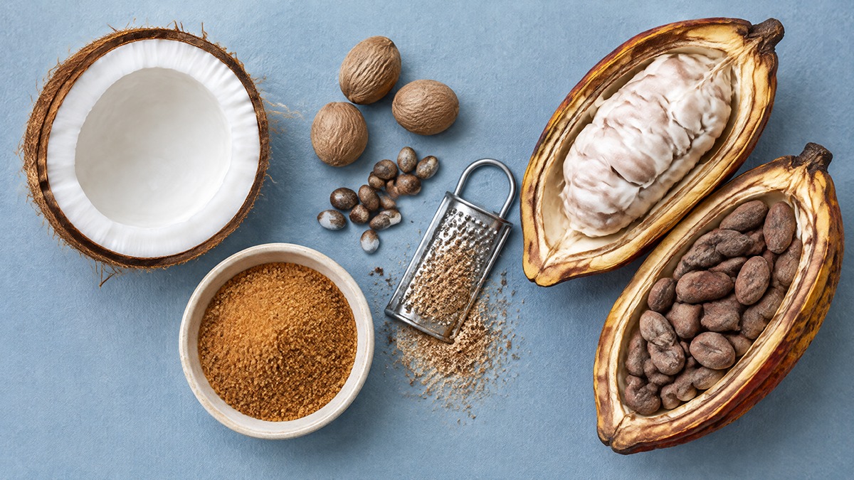 Half a coconut, whole nutmegs, coconut blossom sugar and a split cacao pod with raw cacao beans, seen from above